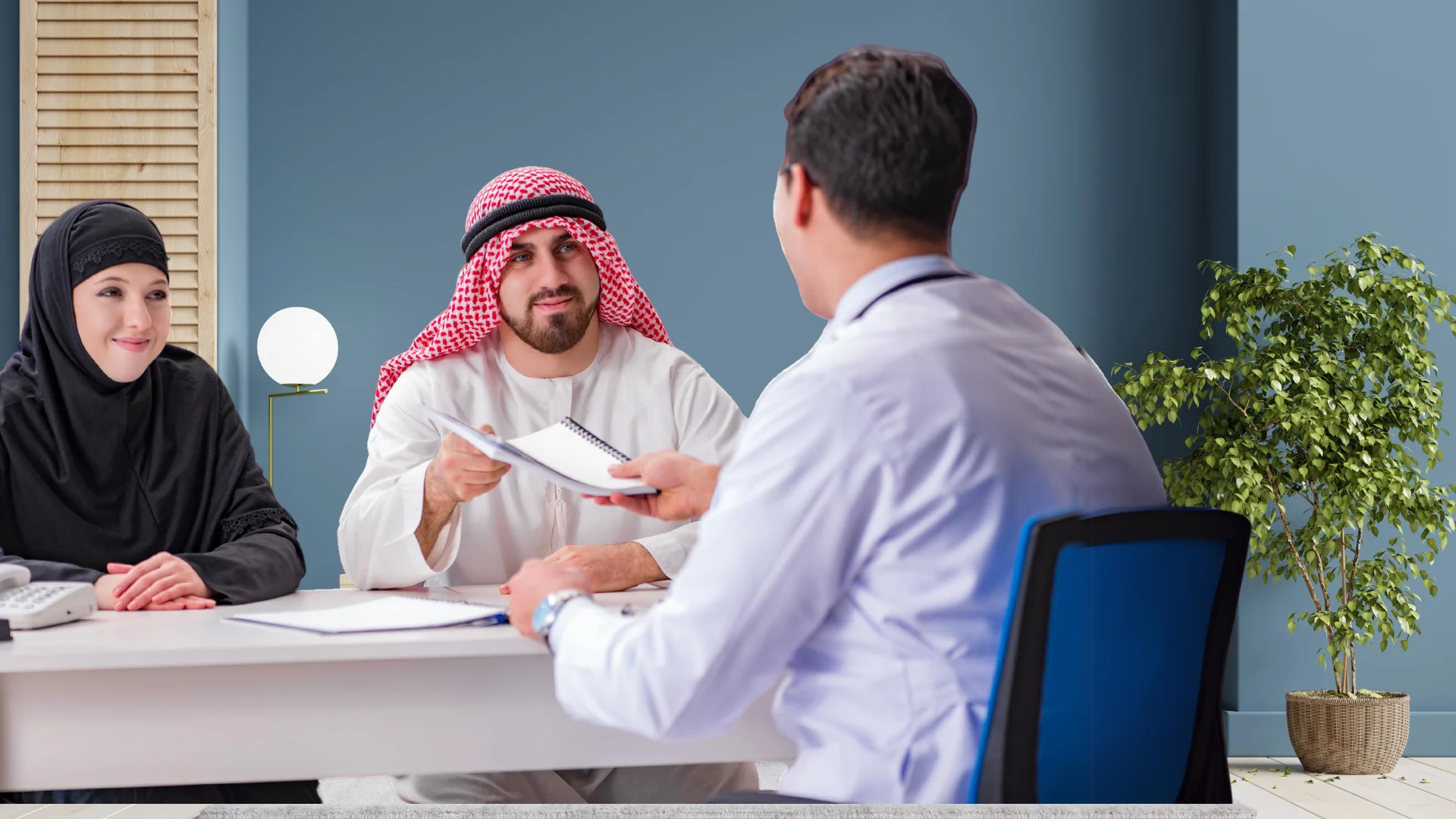 A man and woman in traditional Middle Eastern attire seated across from a male healthcare professional in a clinical office. The man is holding a notepad and pen, possibly discussing a medical consultation with the doctor for Home Healthcare. There's a potted plant and a simple lamp in the room, indicating a comfortable and professional environment.