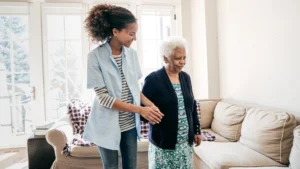 A younger woman assists an elderly woman to walk in a bright, well-lit room with large windows. The younger woman, dressed casually, is guiding the elderly lady, who appears to be frail and is wearing comfortable home clothes. The setting suggests a caring, supportive interaction, likely within a home environment.