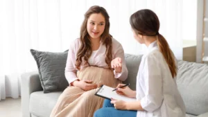 A pregnant woman in a soft pink blouse and cream skirt is conversing with a healthcare professional who is taking notes. They are sitting on a couch in a bright, modern room with a minimalist aesthetic. The scene suggests a prenatal consultation in a relaxed atmosphere.